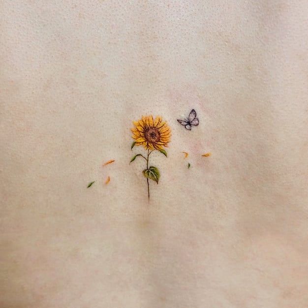 Small, delicate sunflower tattoo with fine details, scattered petals, and a tiny butterfly hovering beside it, creating a soft, minimalist design.