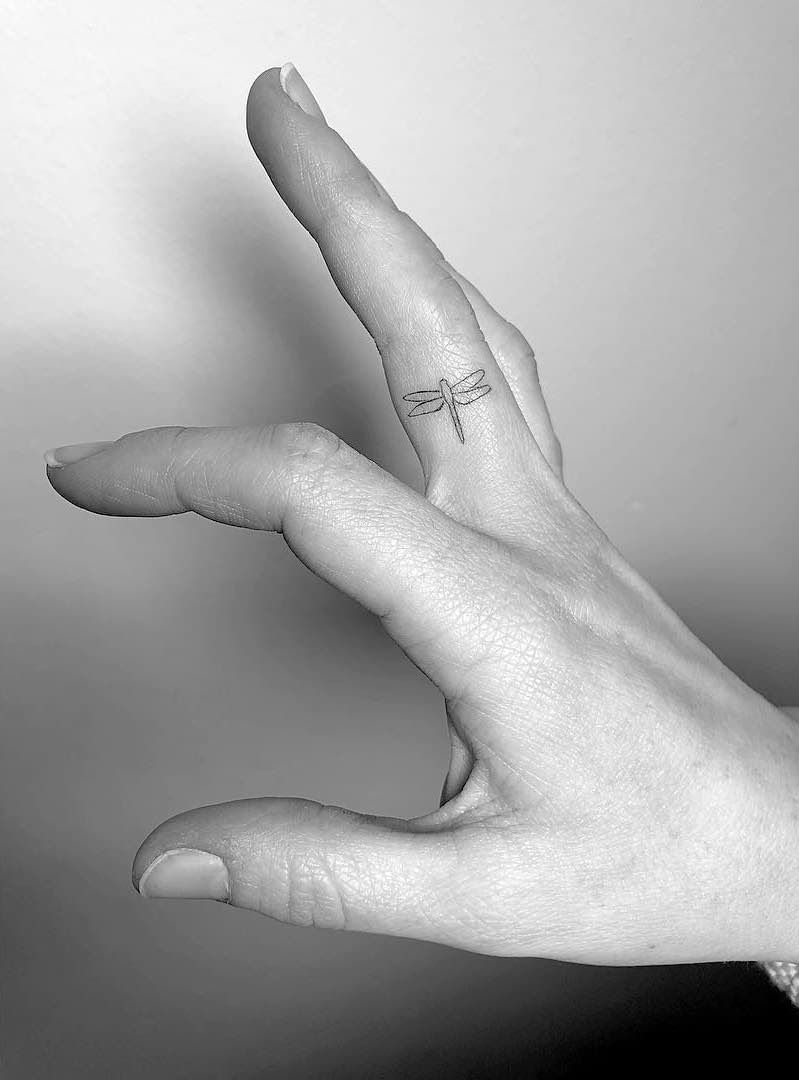 Minimalist dragonfly tattoo on the side of a finger. The fine line design adds a delicate and subtle touch to the hand.
