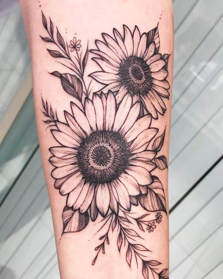 Black and grey forearm tattoo featuring two detailed sunflowers with fine line leaves and floral accents for an elegant botanical design.