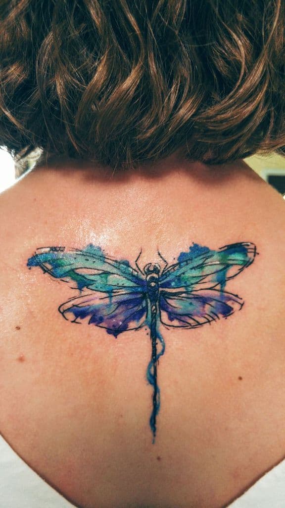 Vibrant watercolor dragonfly tattoo on the upper back, featuring blue and purple hues with an artistic, abstract splash effect.