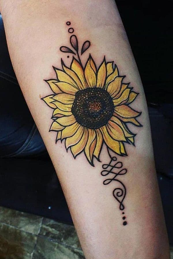 Vibrant sunflower tattoo on forearm with bold black outlines, yellow petals, a dark center, and decorative swirl and dot details.