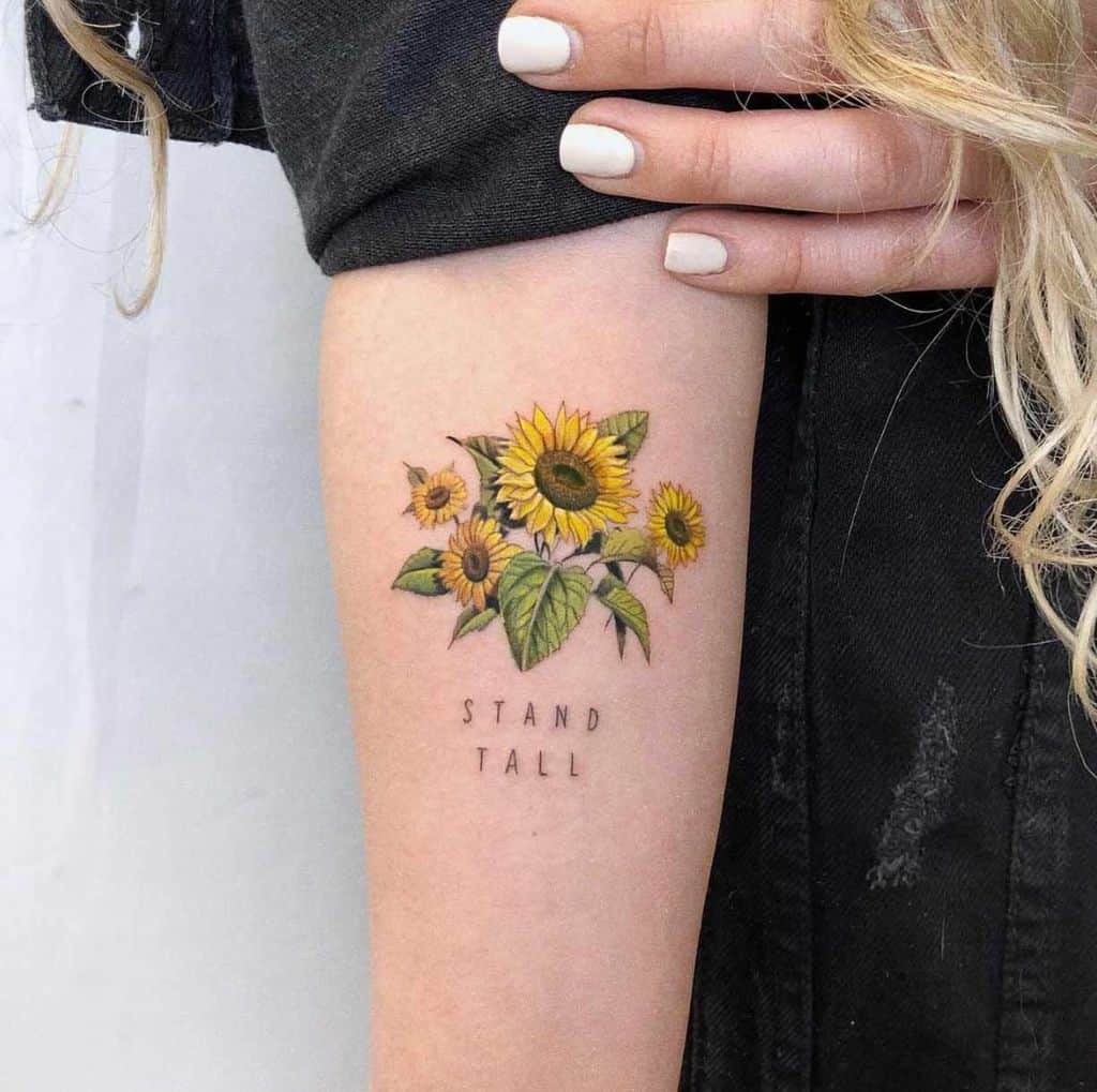 Small sunflower bouquet tattoo on upper arm with fine details, vibrant yellow petals, green leaves, and 'STAND TALL' text below.