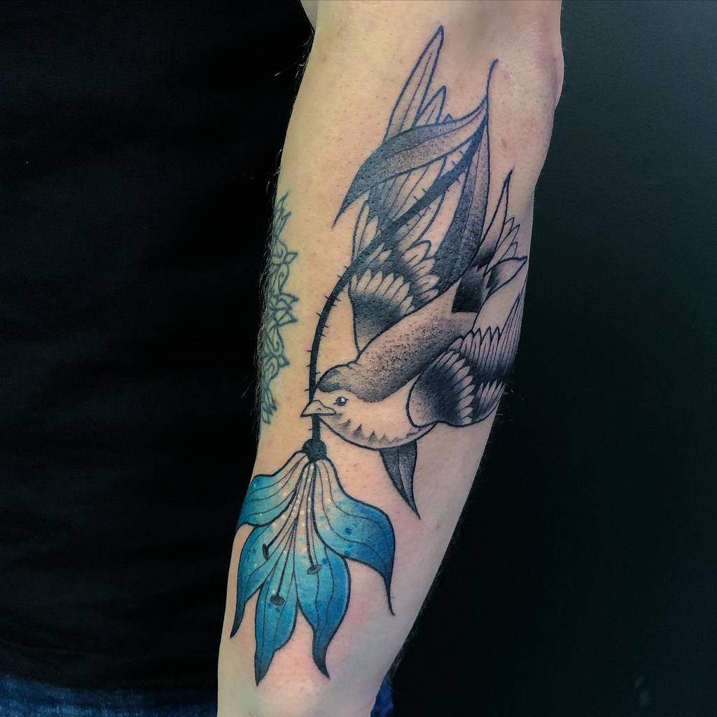 Tattoo of a swallow holding a blue lily, blending traditional and neo-traditional styles with fine details and bold shading