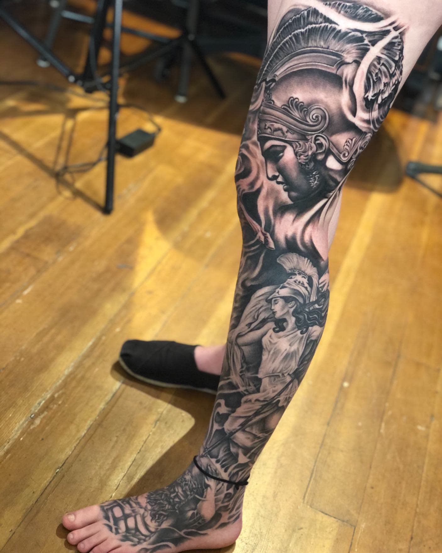 A leg adorned with intricate warrior-themed tattoos in black and grey, extending from the foot to the thigh