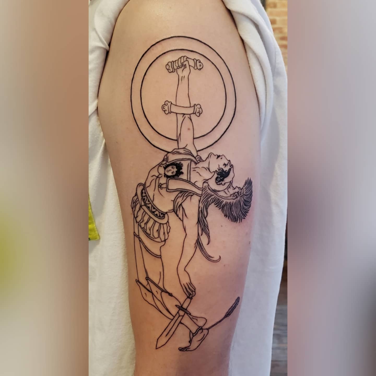 Tattoo on upper arm depicting a hanging figure with swords and circular elements