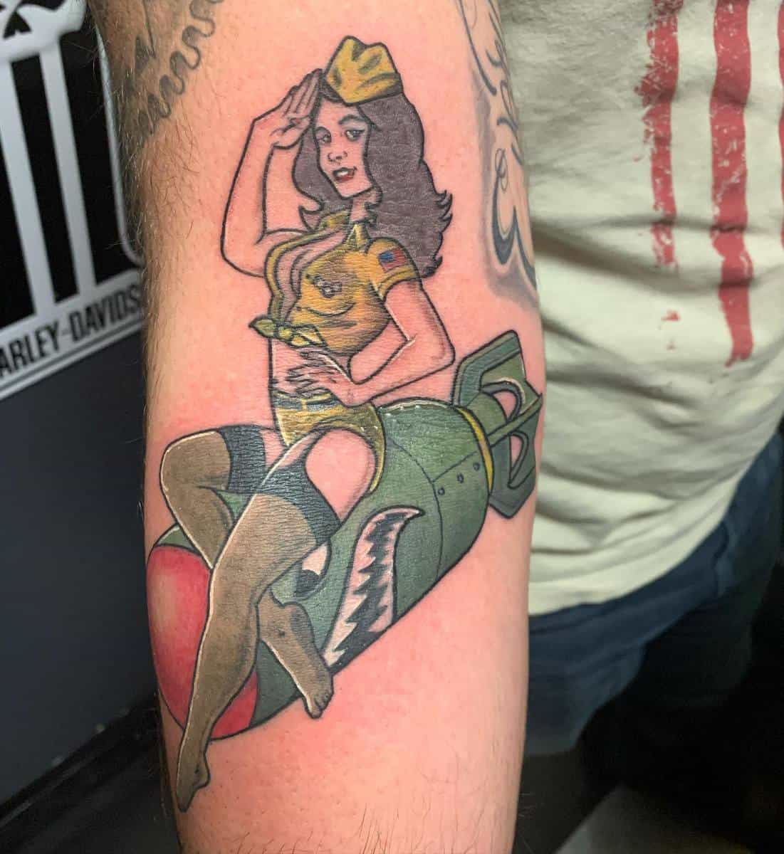 Tattoo of a pin-up girl saluting while sitting on a bomb with shark teeth, located on an arm