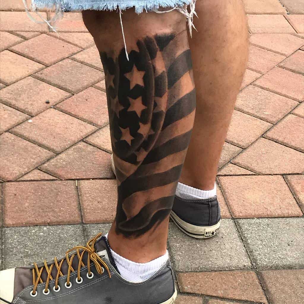 Dynamic black and grey American flag calf tattoo with a bold waving effect, showcasing stars and stripes in a striking patriotic design