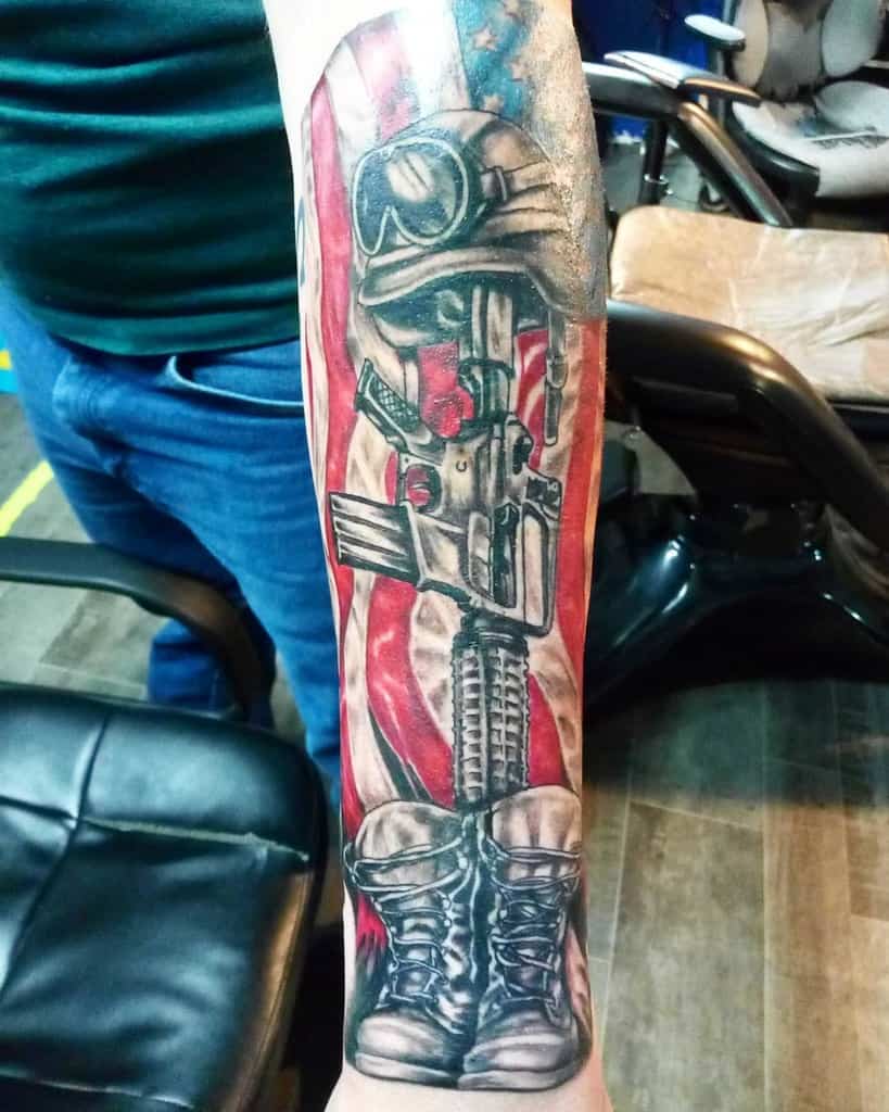 Tattoo of a soldier's helmet, rifle, and boots with an American flag background on an arm