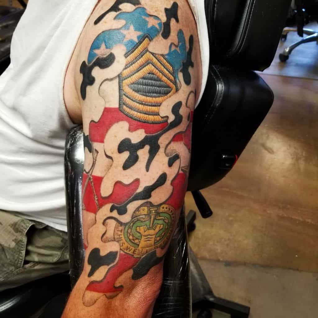 Bold sleeve tattoo featuring a camouflage pattern integrated with the American flag, military insignia, and detailed patches to honor service and patriotism