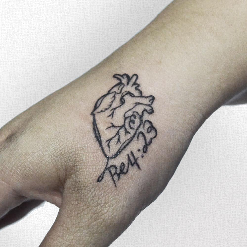 A minimalistic anatomical heart tattoo on the hand with "Be 4:23" inscribed