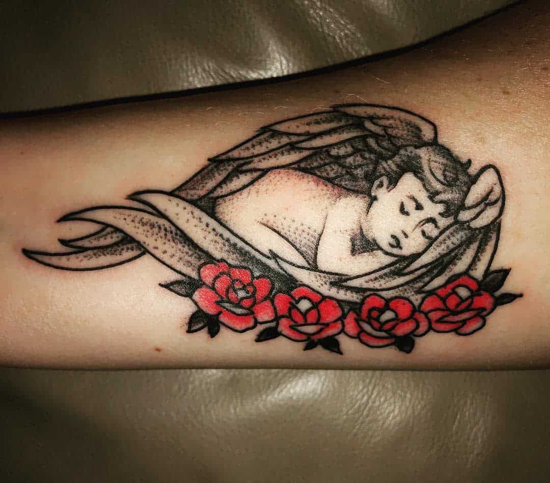 Tattoo of a sleeping baby with wings resting on red roses inked on an arm