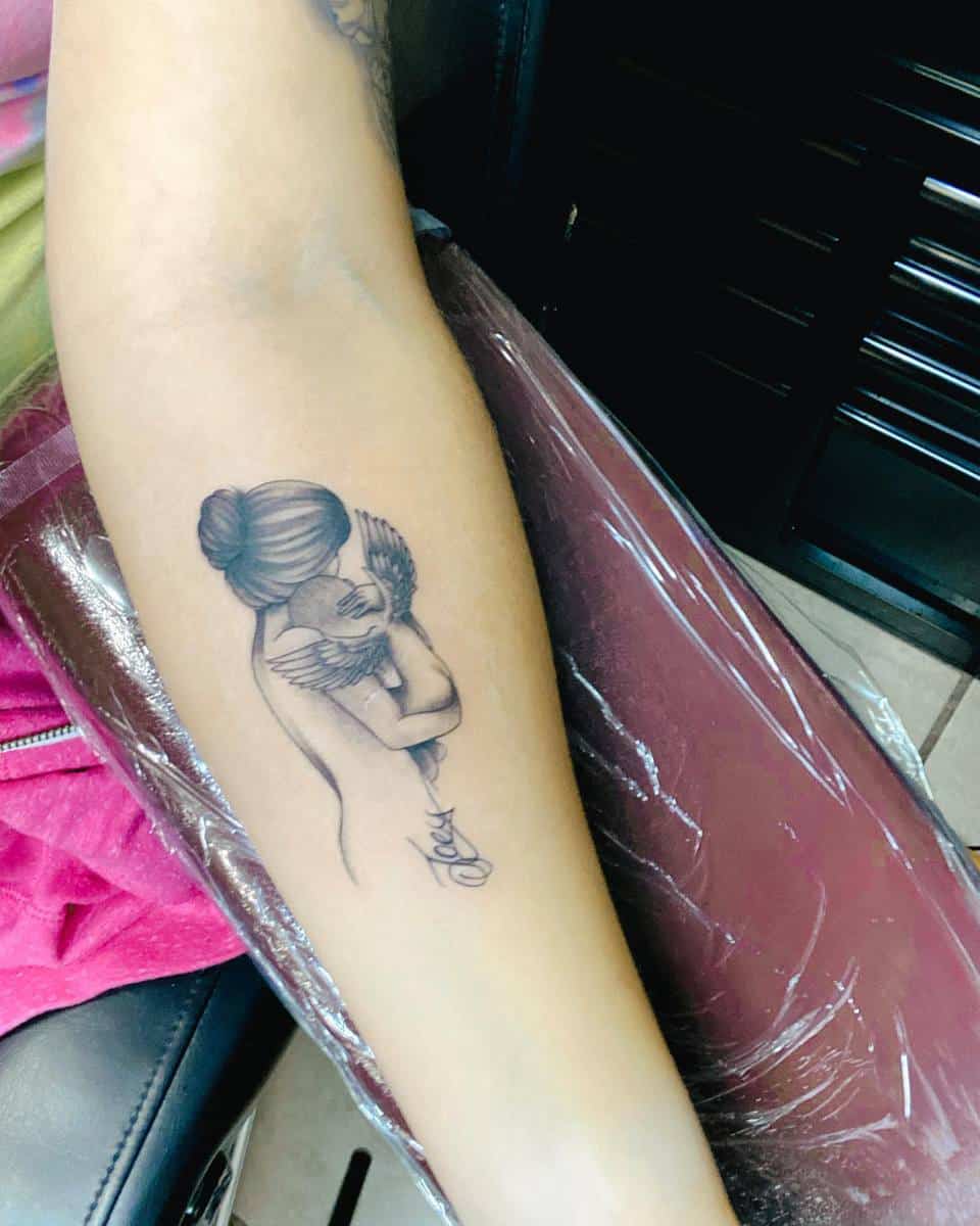 Forearm tattoo of a woman with a bun holding a winged baby, with a name inscribed below