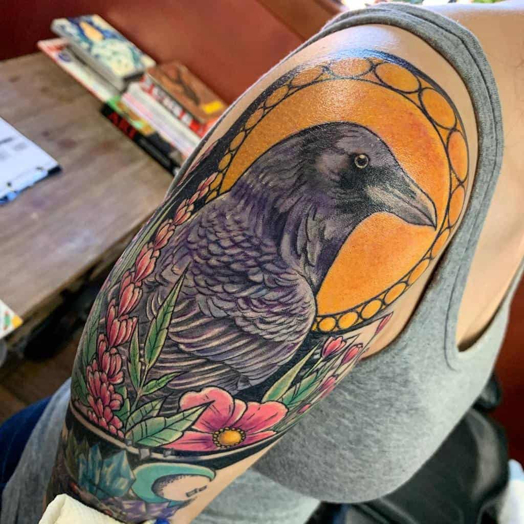Art Nouveau-style tattoo of a raven framed by a golden circular pattern, surrounded by lush pink flowers and greenery with crystals and moon below