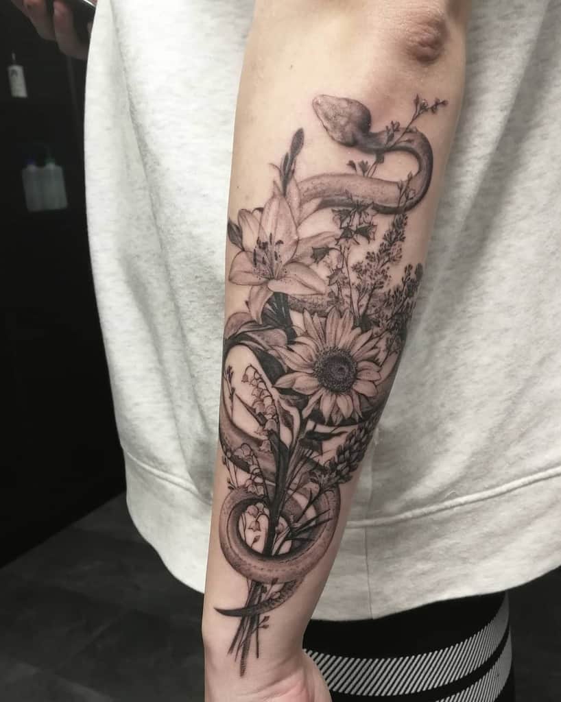 Black and grey tattoo of a snake weaving through lilies, sunflowers, and wildflowers