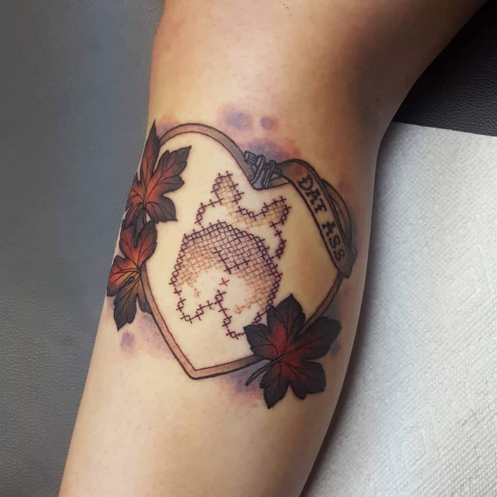 Creative cross-stitch style tattoo of a corgi butt inside a heart-shaped frame with autumn leaves and a banner reading 'DAT ASS'