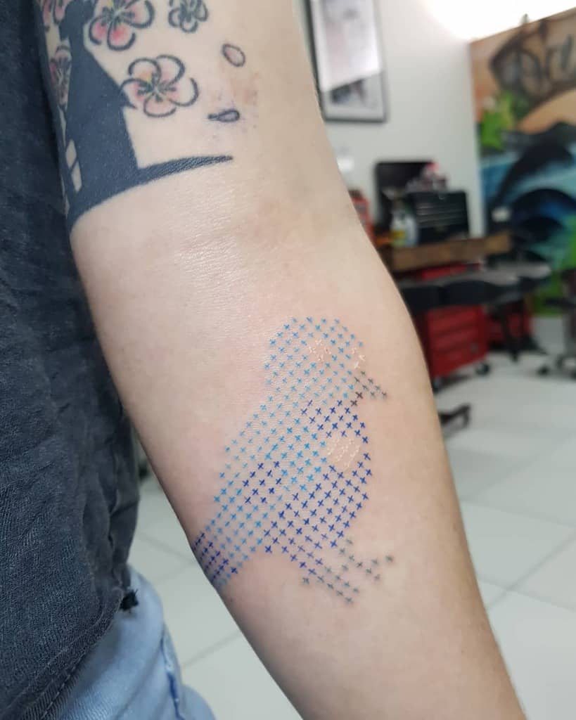 Minimalist cross-stitch style tattoo of a bird in shades of blue, creating a pixelated embroidery effect