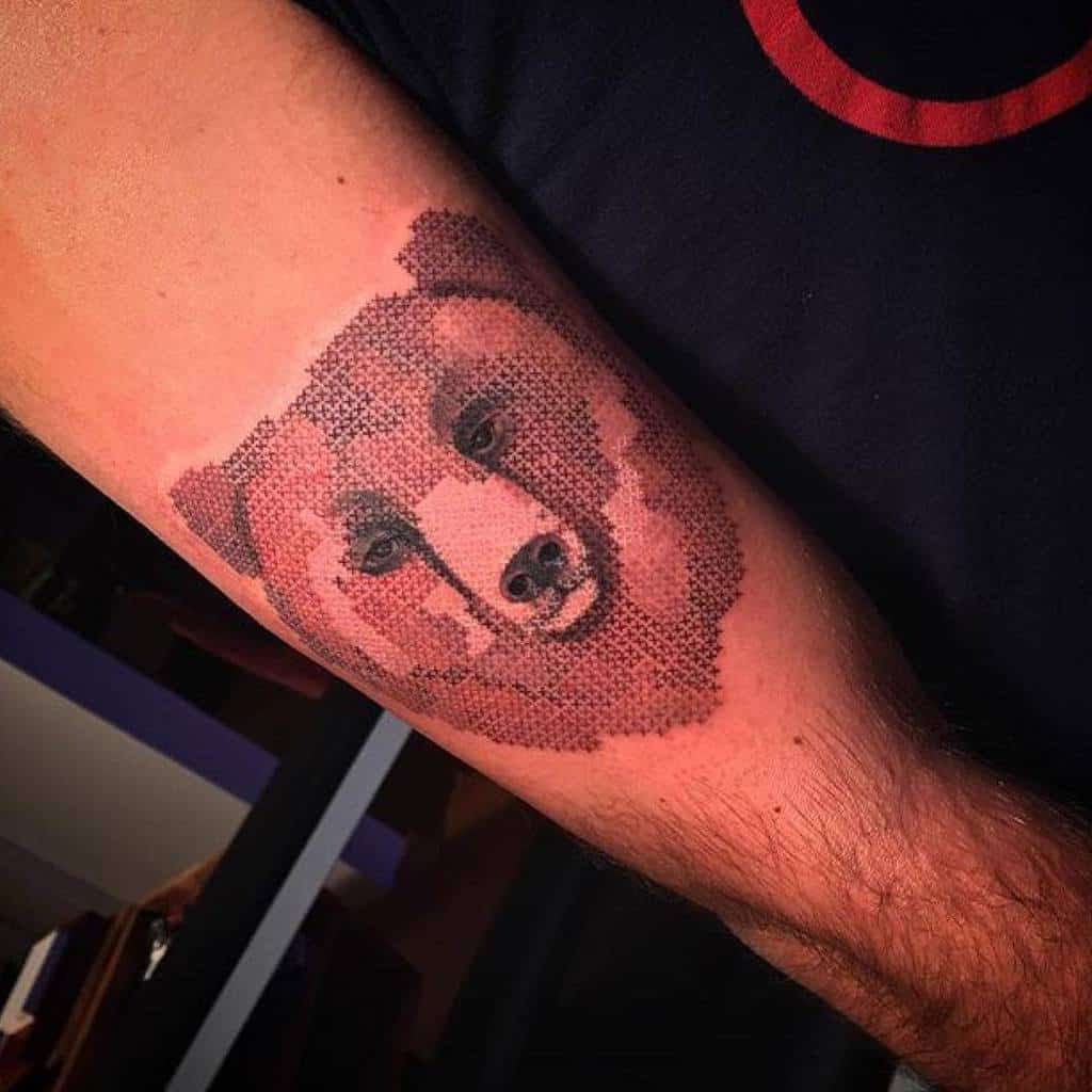 Intricate cross-stitch style tattoo of a bear’s face, creating a pixelated embroidery effect with warm brown tones