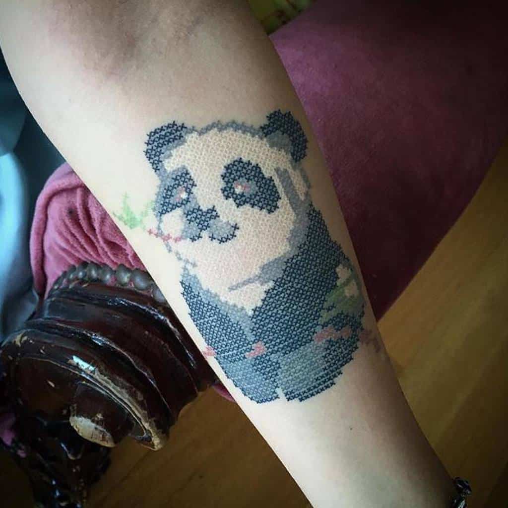 Unique cross-stitch style tattoo of a smiling panda holding bamboo, creating a pixelated embroidery effect