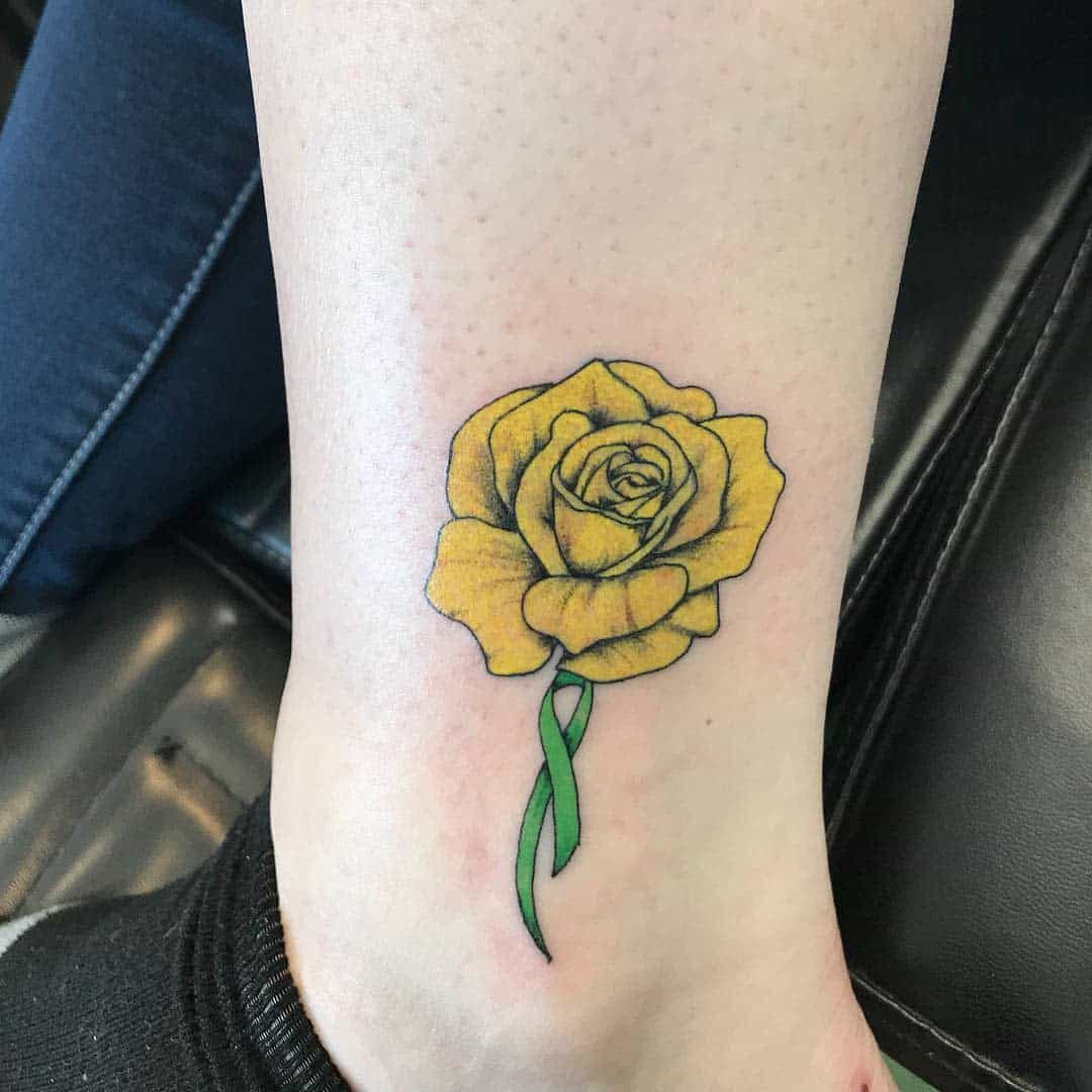 A yellow rose tattoo with a green stem on the lower leg above the ankle