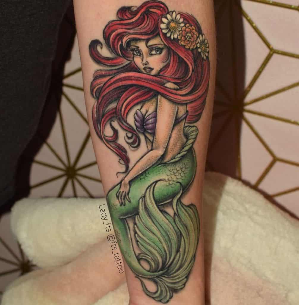 A stunning tattoo of Ariel, styled with flowing red hair, floral crown, and shimmering green tail, showcasing her iconic mermaid beauty