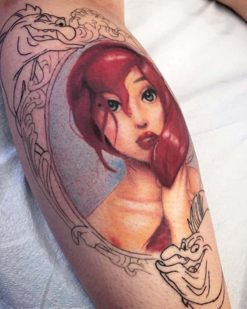 A delicate tattoo of Ariel framed in a decorative outline, showcasing her soft red hair, serene expression, and vivid green eyes
