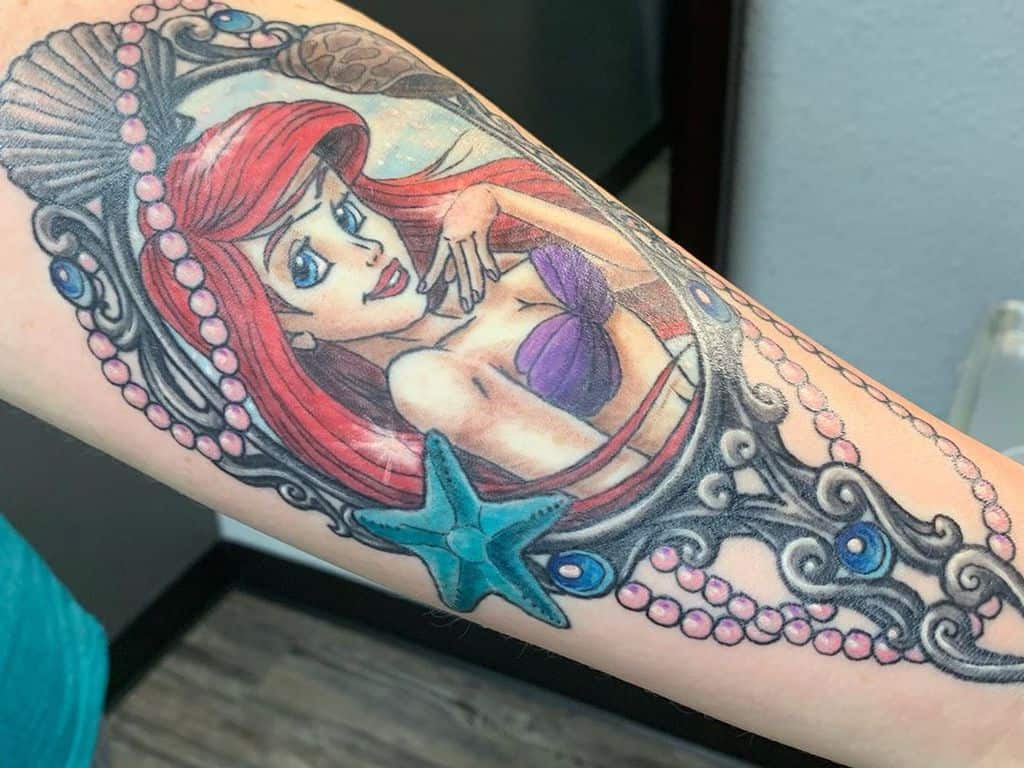 A colorful tattoo of Ariel framed by ornate pearls, seashells, and a starfish, highlighting her signature red hair and purple top