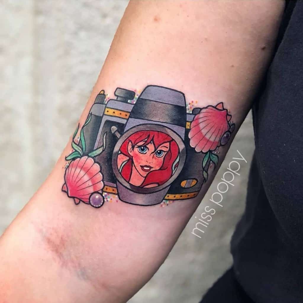 A creative tattoo featuring Ariel's face framed inside a vintage camera lens, surrounded by seashells and floral accents