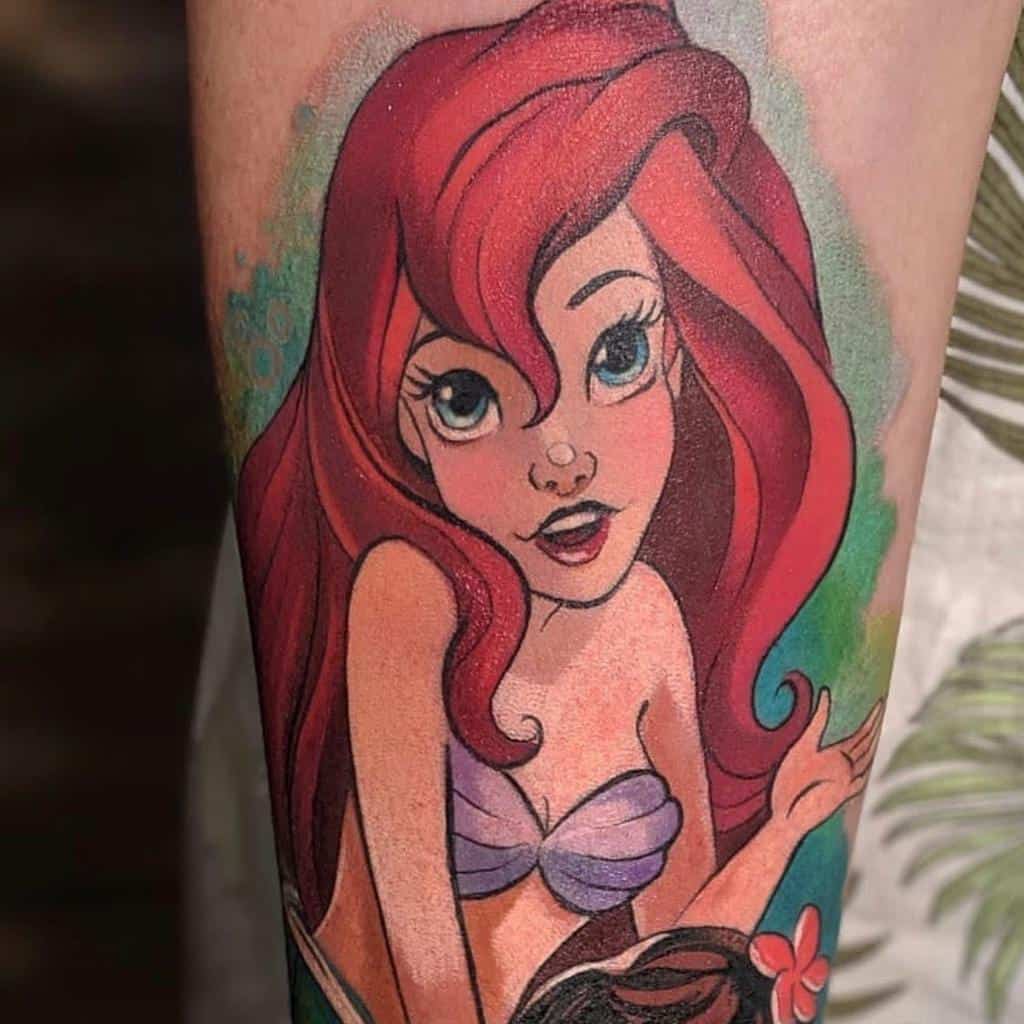 A vibrant tattoo of Ariel from The Little Mermaid, highlighting her flowing red hair, purple seashell top, and expressive, curious gaze