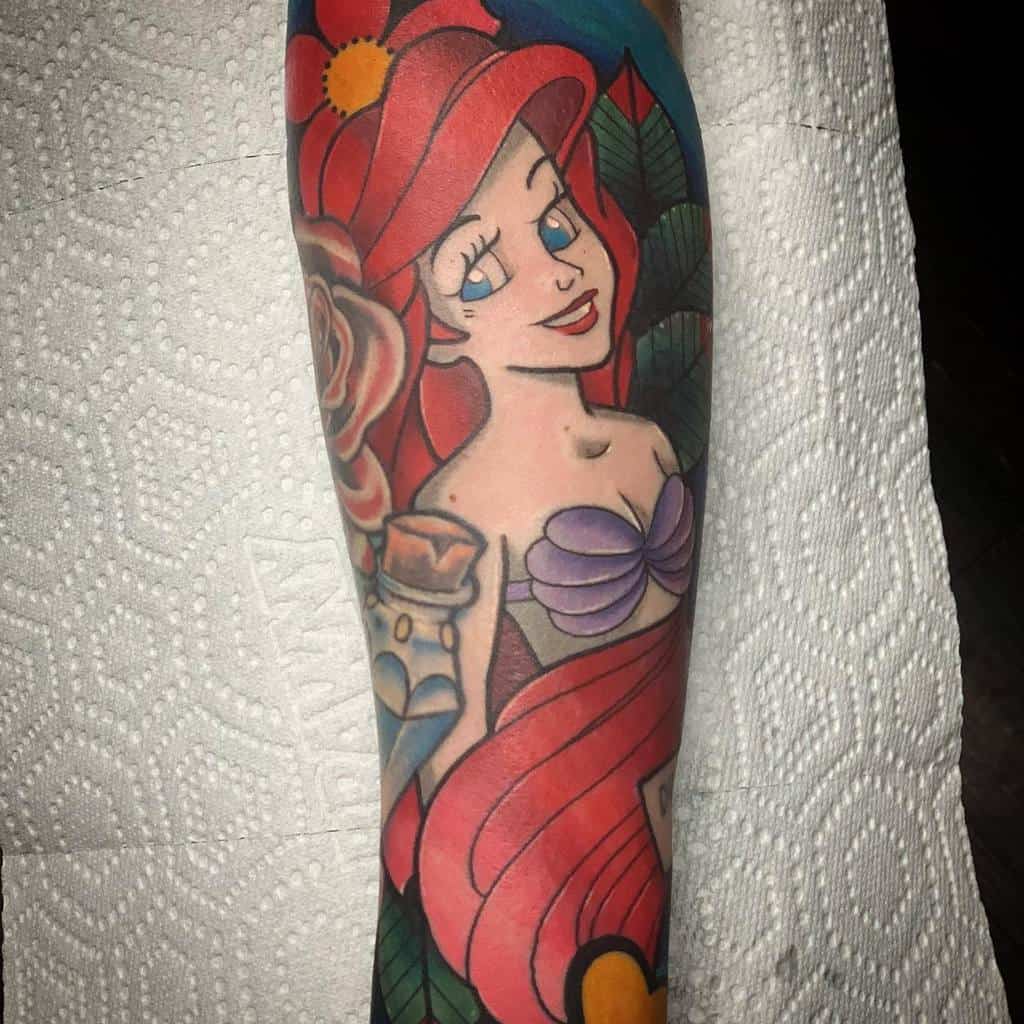 A colorful tattoo of Ariel featuring her red hair, purple seashell top, surrounded by a rose, vibrant leaves, and a nautical theme