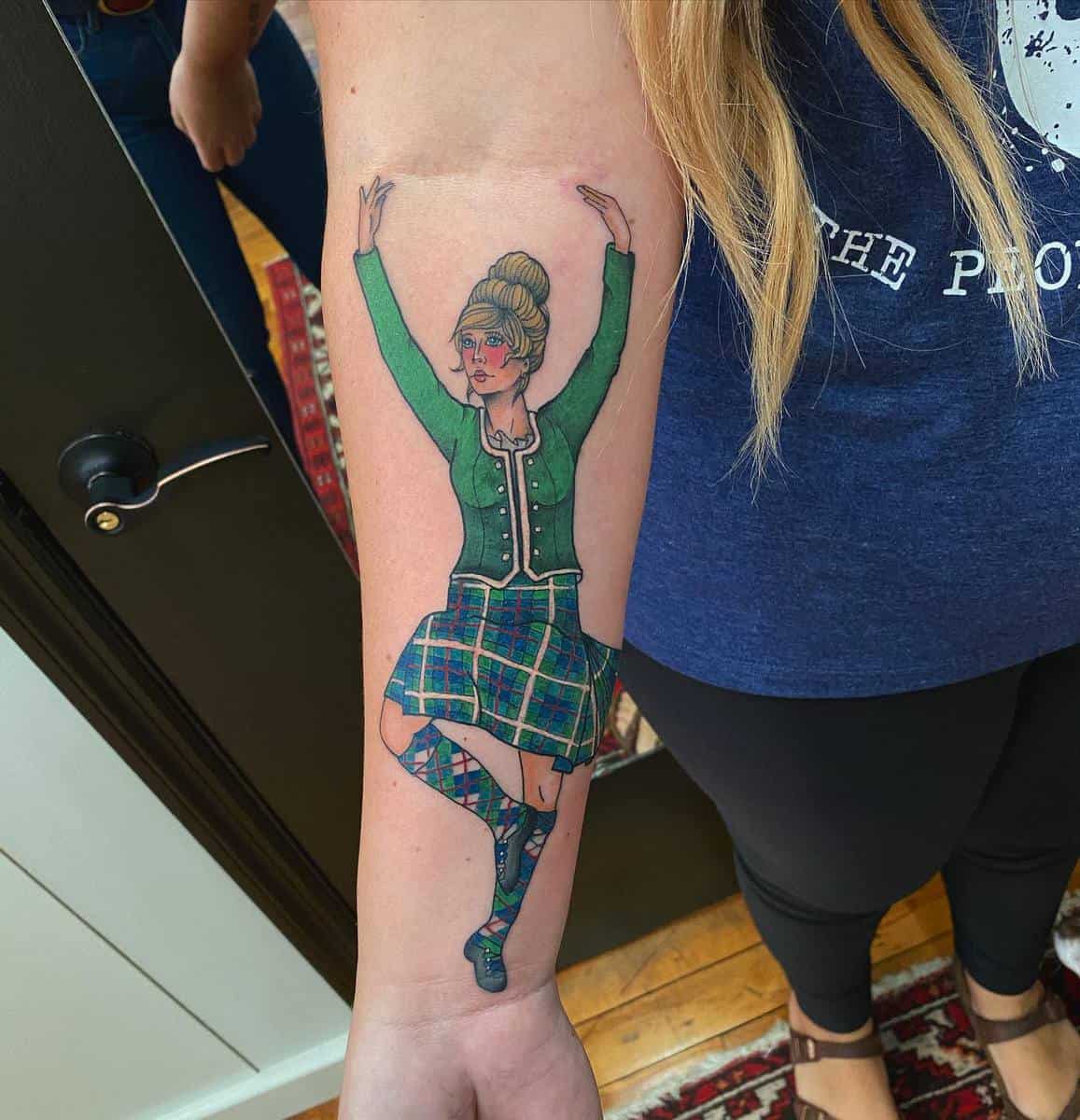 Forearm tattoo of a dancer wearing traditional Scottish attire including a tartan skirt and green jacket