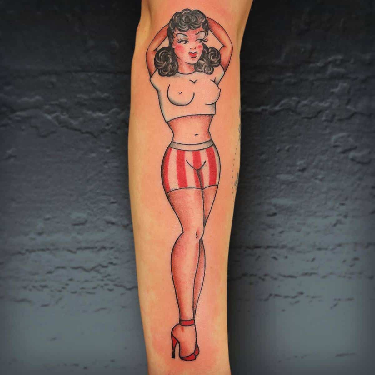 Forearm tattoo of a pin-up girl with long hair, wearing red-striped shorts and red heels