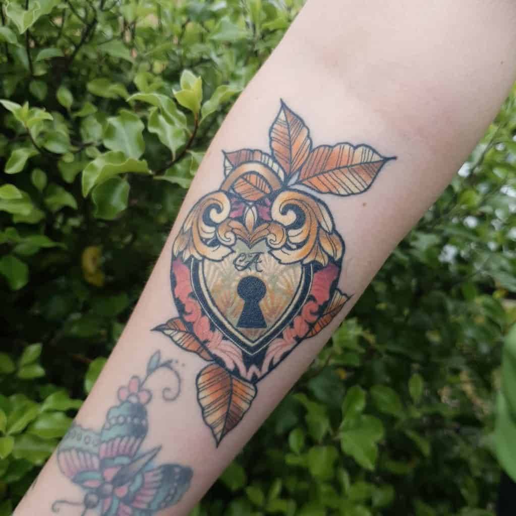 Art Nouveau-style tattoo of an ornate heart-shaped lock with intricate golden details, swirling patterns, and autumn leaves