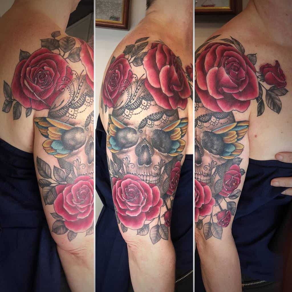 A vibrant shoulder tattoo featuring a lace-detailed skull, colorful wings, and bold red roses, blending beauty with mortality