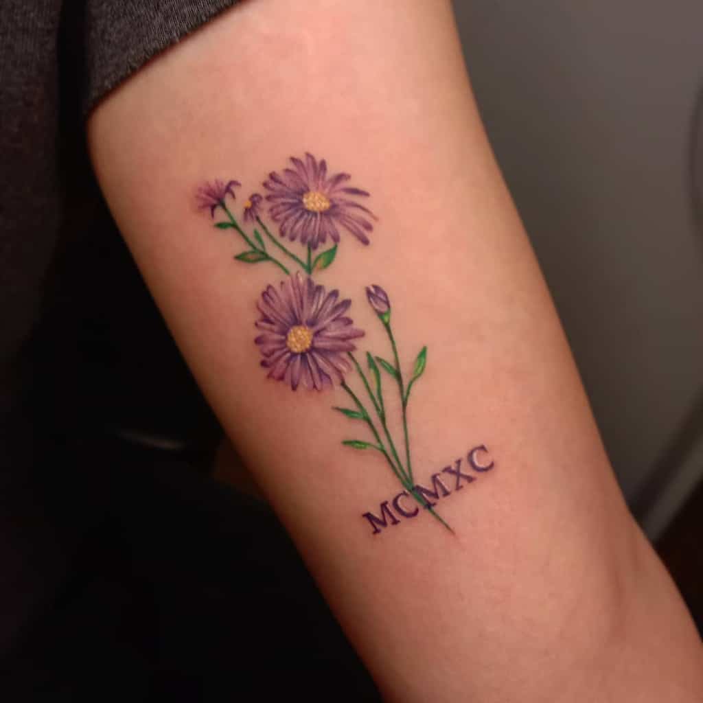 Tattoo of purple aster flowers and green leaves with the Roman numerals MCMXC on an arm