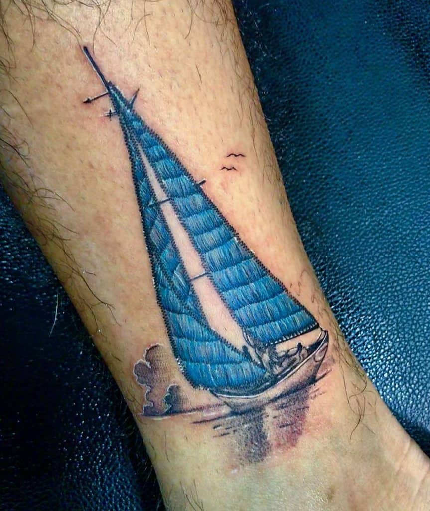 Embroidery-style tattoo of a blue sailboat on a person's leg, with detailed stitching effects, ocean waves, and birds in the sky.