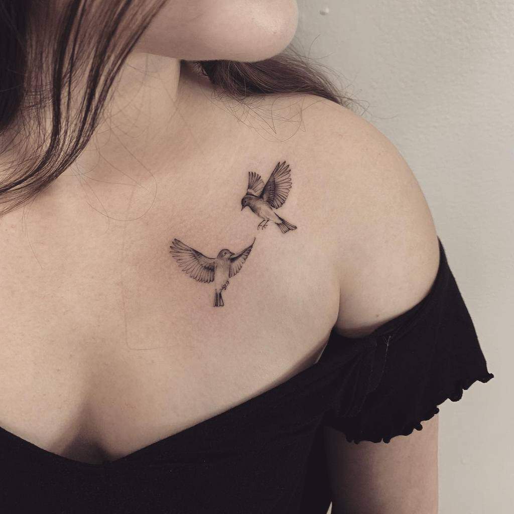Two small single needle bird tattoos on woman's upper chest above off-shoulder black top