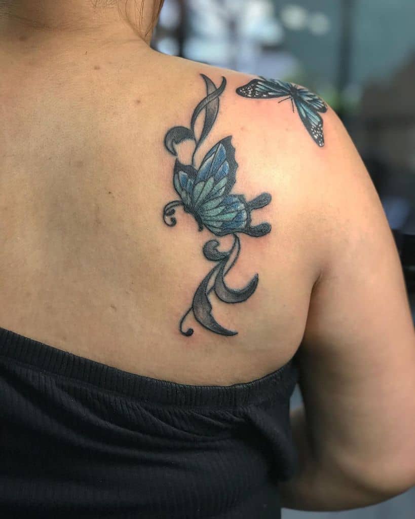 Blue butterfly tattoos on a person's upper back featuring striking blue and black coloring with intricate vine-like details