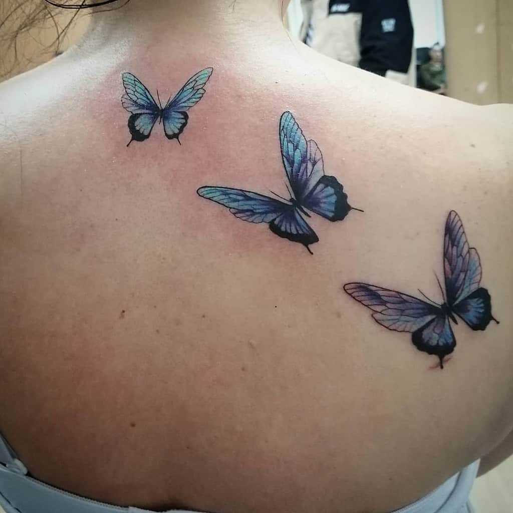 Three striking blue butterfly tattoos adorn a person's upper back, blending shades of blue and purple
