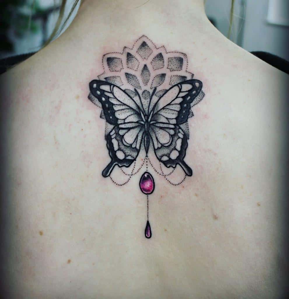 Back tattoo of a butterfly with an ornamental design and a dangling gem at the bottom
