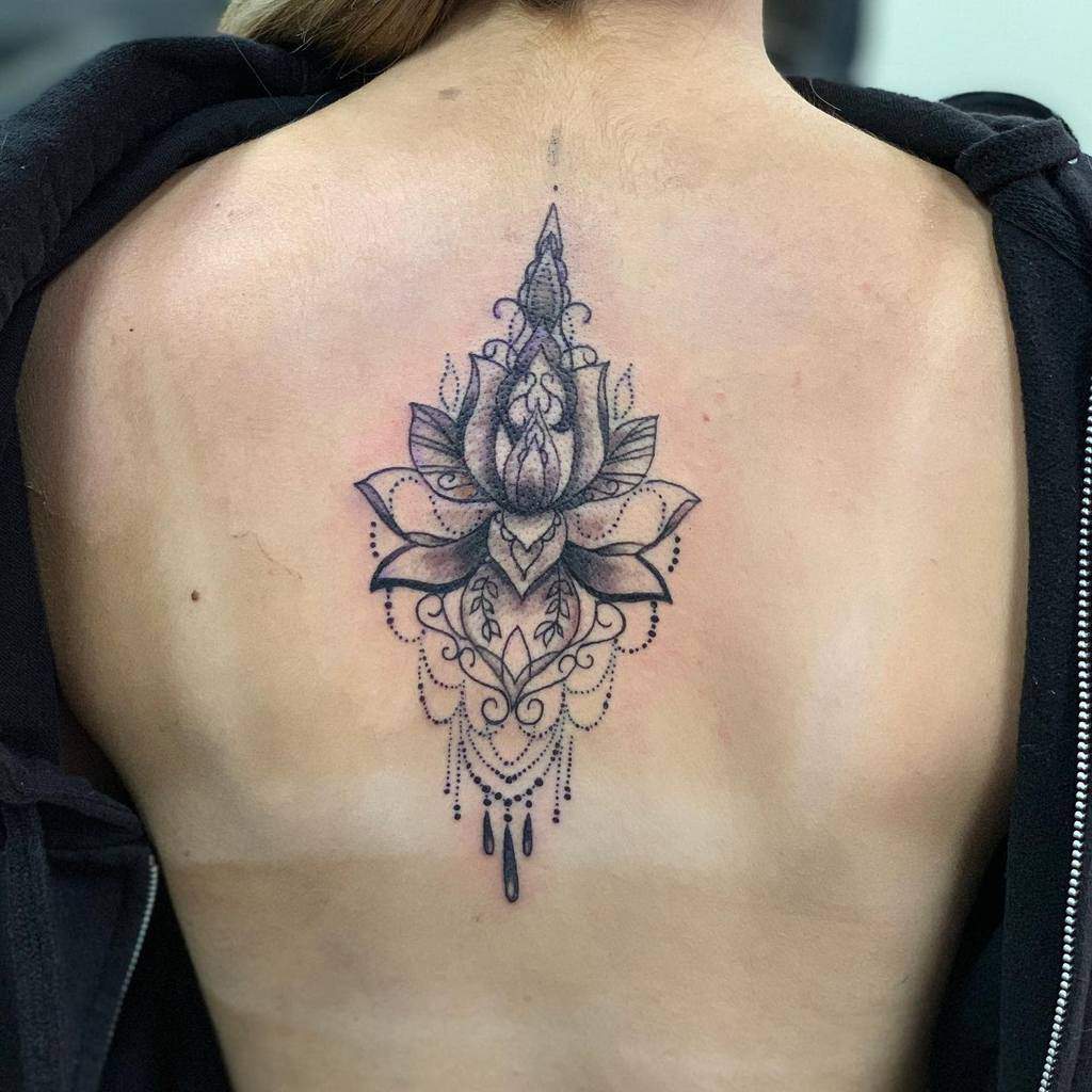 Black ink back tattoo featuring a detailed lotus flower with intricate mandala patterns, chandelier-style beads, and ornamental flourishes