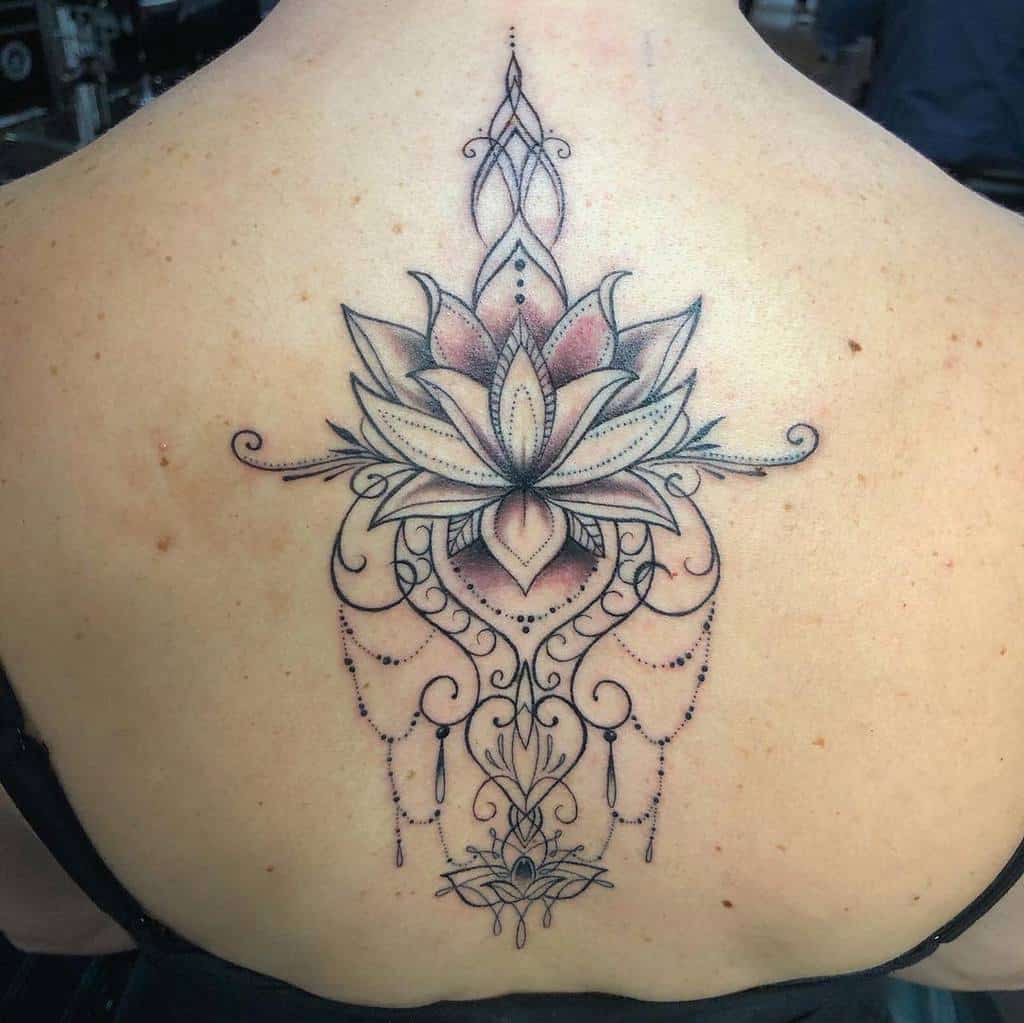 Ornate black and shaded ink back tattoo featuring a detailed lotus flower with mandala elements, a subtle heart shape, chandelier-style beads, and swirling patterns