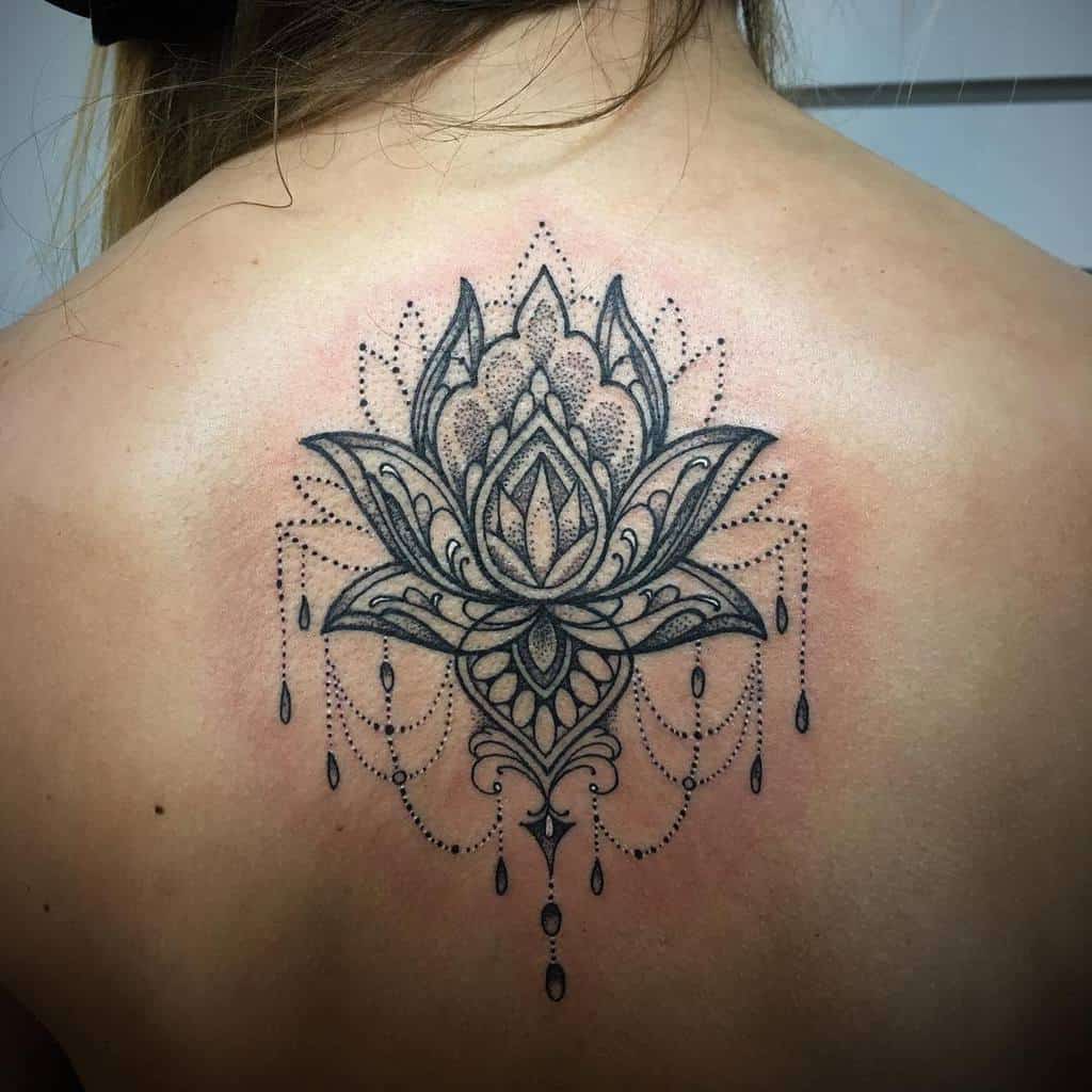 Elegant black ink back tattoo featuring a detailed lotus flower with intricate dot work, mandala patterns, chandelier-style beads, and teardrop accents