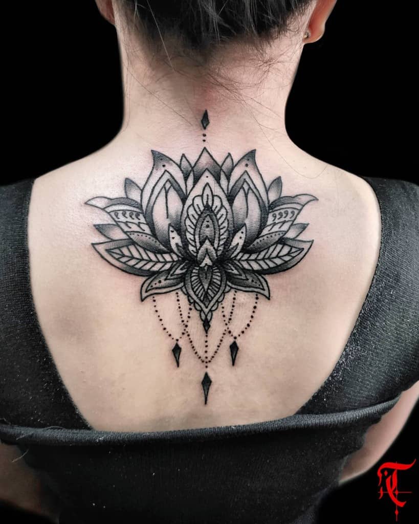 Bold black ink tattoo featuring a detailed lotus flower with intricate geometric patterns, subtle shading, and chandelier-style beadwork