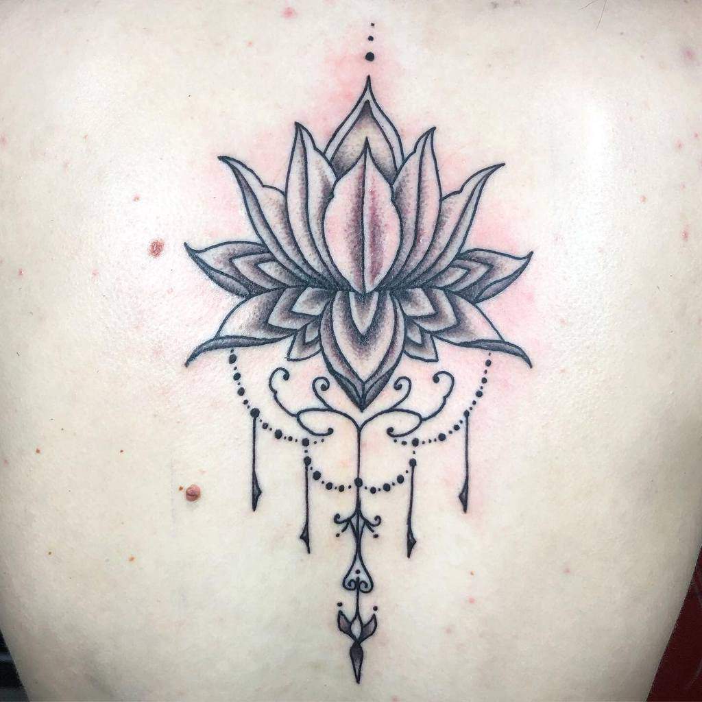 Delicate black and grey ink back tattoo featuring a shaded lotus flower with intricate line work, chandelier-style beads, and ornamental flourishes