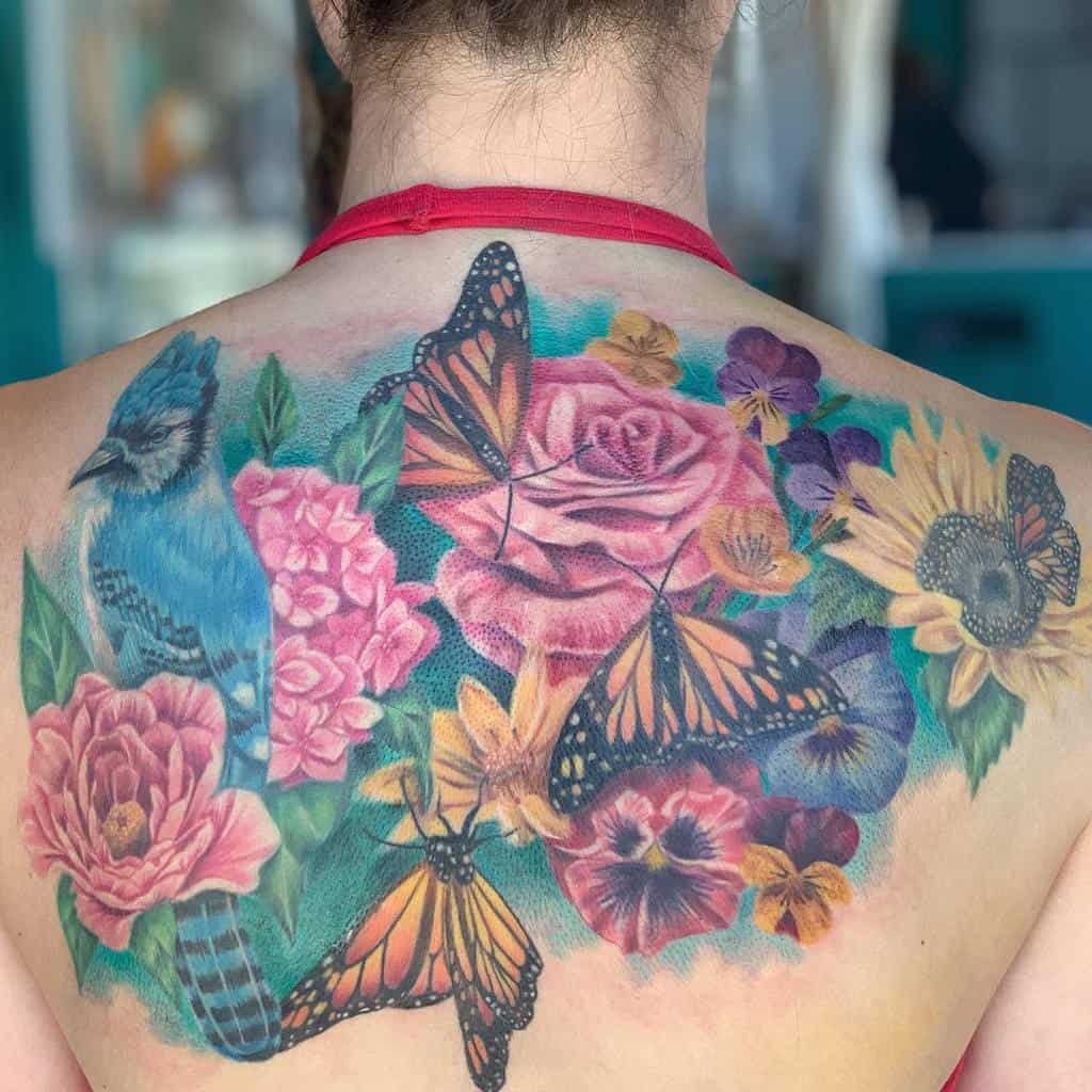 A stunning back tattoo featuring vibrant flowers like sunflowers, roses, and peonies, accompanied by a graceful blue jay and enchanting monarch butterflies