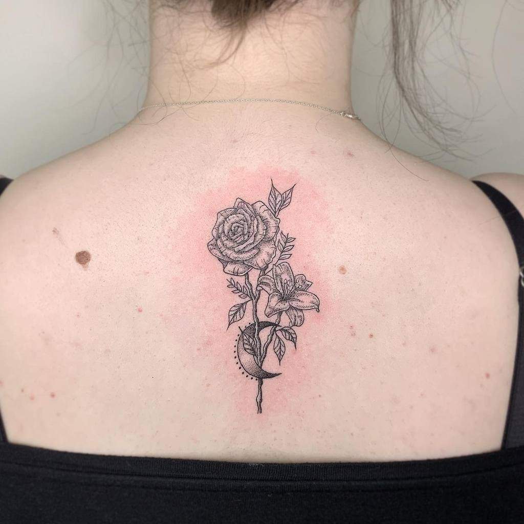 Delicate black and grey back tattoo of a rose and lily intertwined with a crescent moon, symbolizing beauty, balance, and transformation