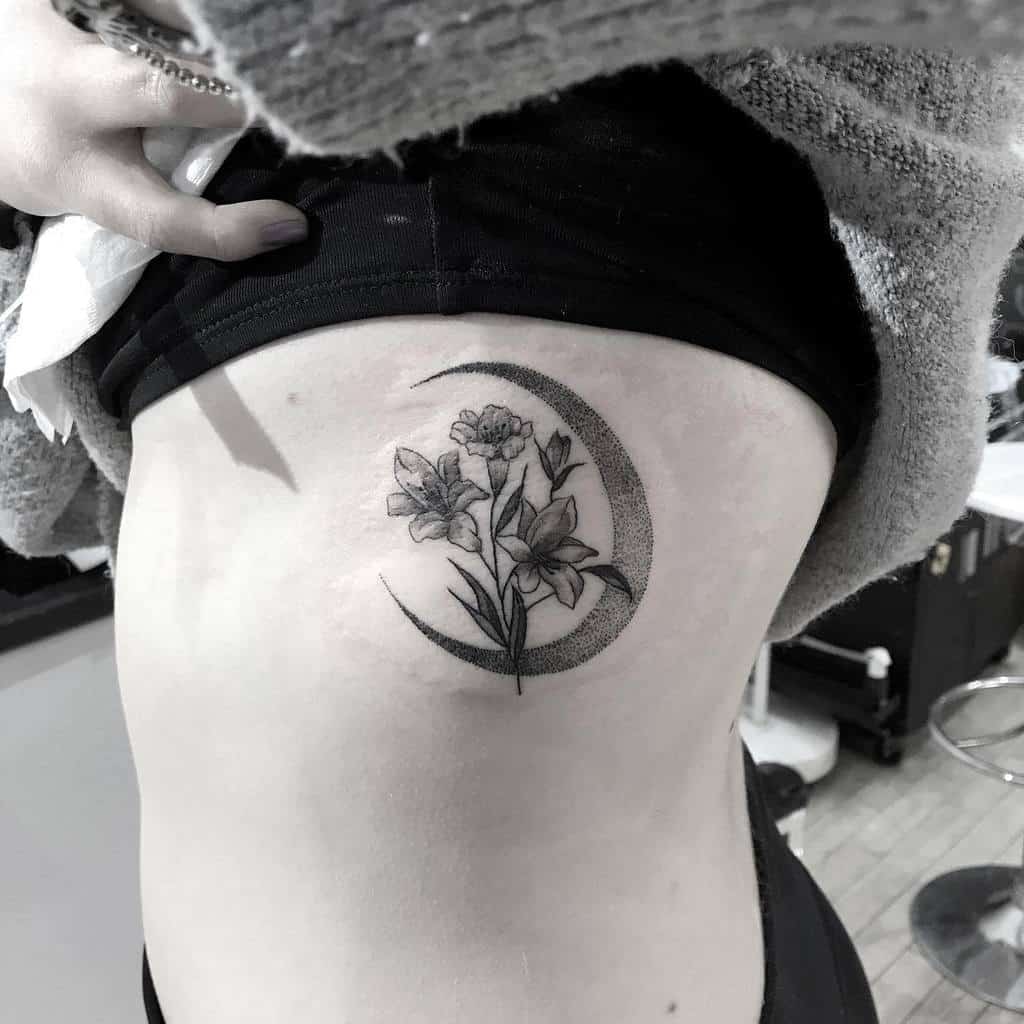 Elegant black and grey tattoo of lilies and wildflowers within a dotted crescent moon, symbolizing nature, femininity, and mysticism