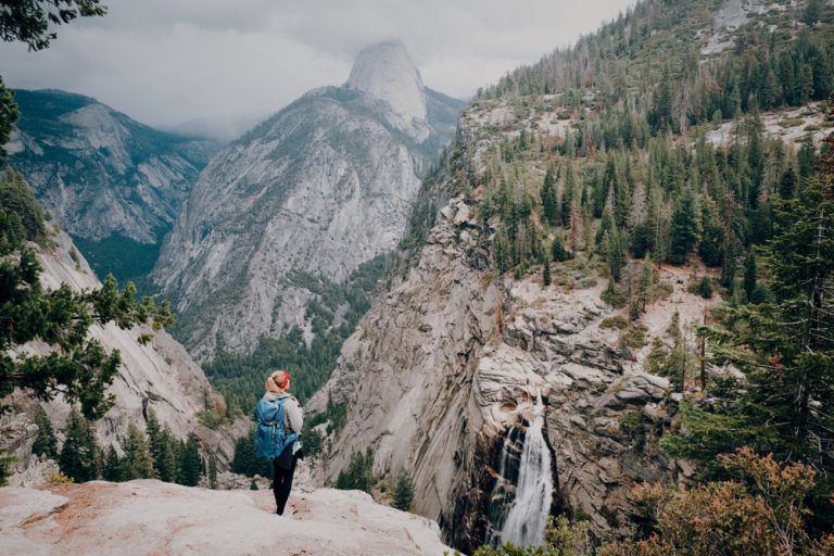 A hiker stands on a cliff, enjoying backpacking comfort as they gaze at a tall waterfall cascading into a forested valley with distant mountains