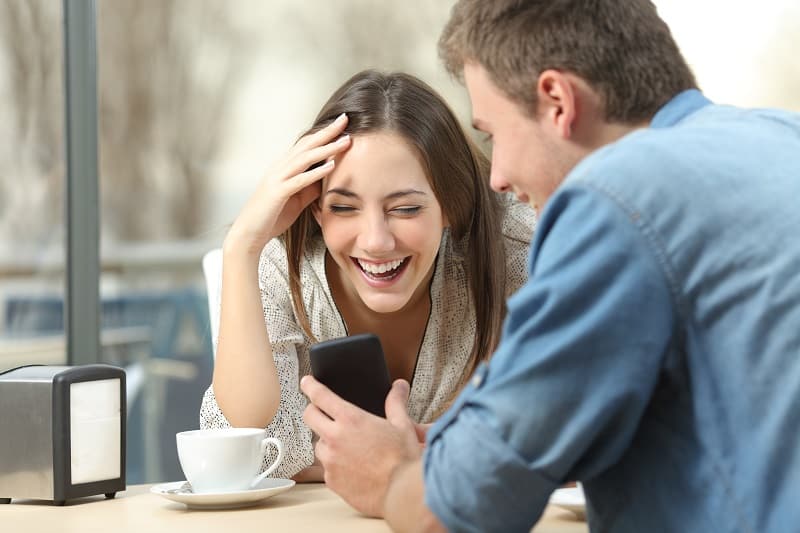 Cheerful,Couple,Laughing,Watching,Media,In,Smart,Phone,Sitting,In