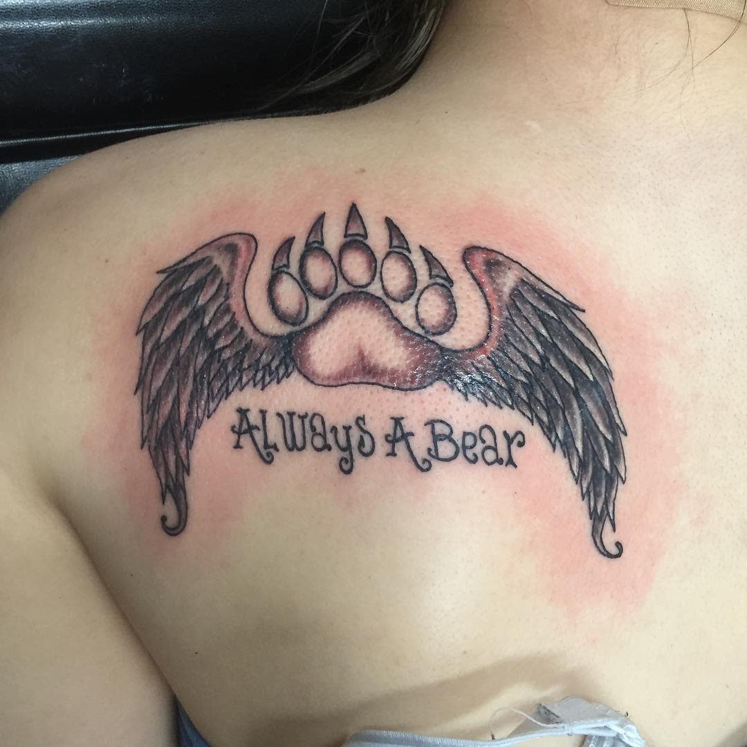 Tattoo of a bear paw with wings and the text "ALWAYS A BEAR" on a person's upper back